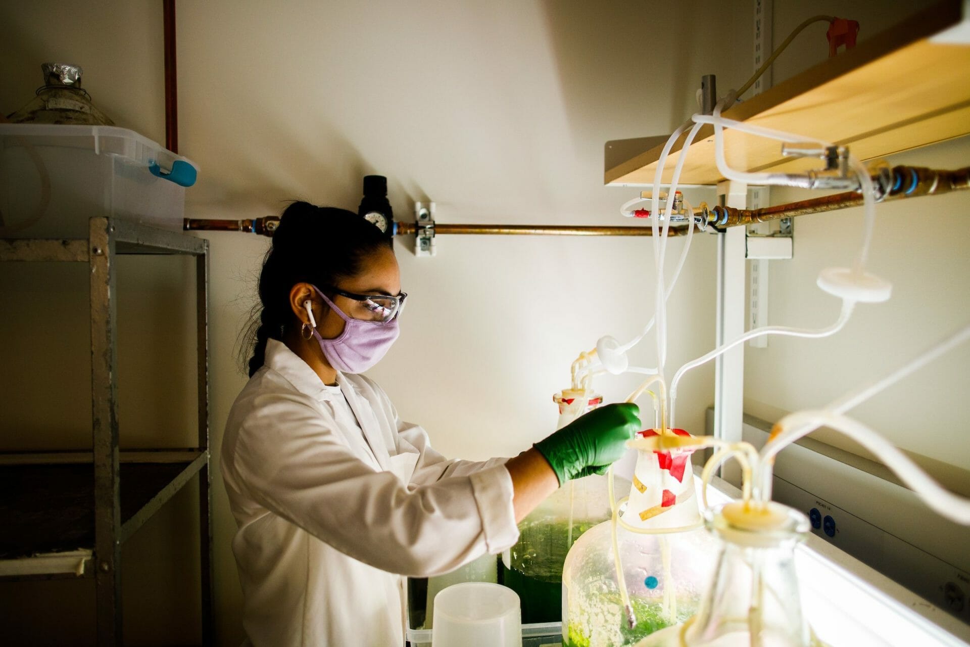 Woman in lab wearing lab coat, gloves and mask conducting experiment.